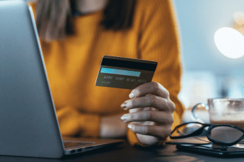 A woman in a yellow jumper sitting at her laptop. She holds her payment card in her hand.