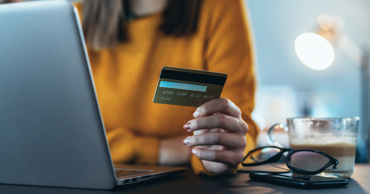 A woman in a yellow jumper sitting at her laptop. She holds her payment card in her hand.